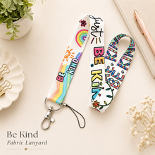 Load image into Gallery viewer, Be Kind fabric lanyard with colourful rainbow and positive message design, silver clasp and keyring, styled on a neutral desk flat lay background.

