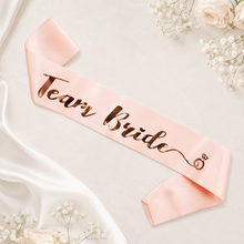 Load image into Gallery viewer, Pink Team Bride satin sash with gold lettering, styled as a bridal party accessory for hens parties and bridesmaid gift bags.
