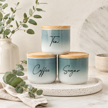 Load image into Gallery viewer, Three ceramic canisters with bamboo lids labeled Tea, Coffee, and Sugar, stacked on a round tray with eucalyptus branches in a styled kitchen setting.
